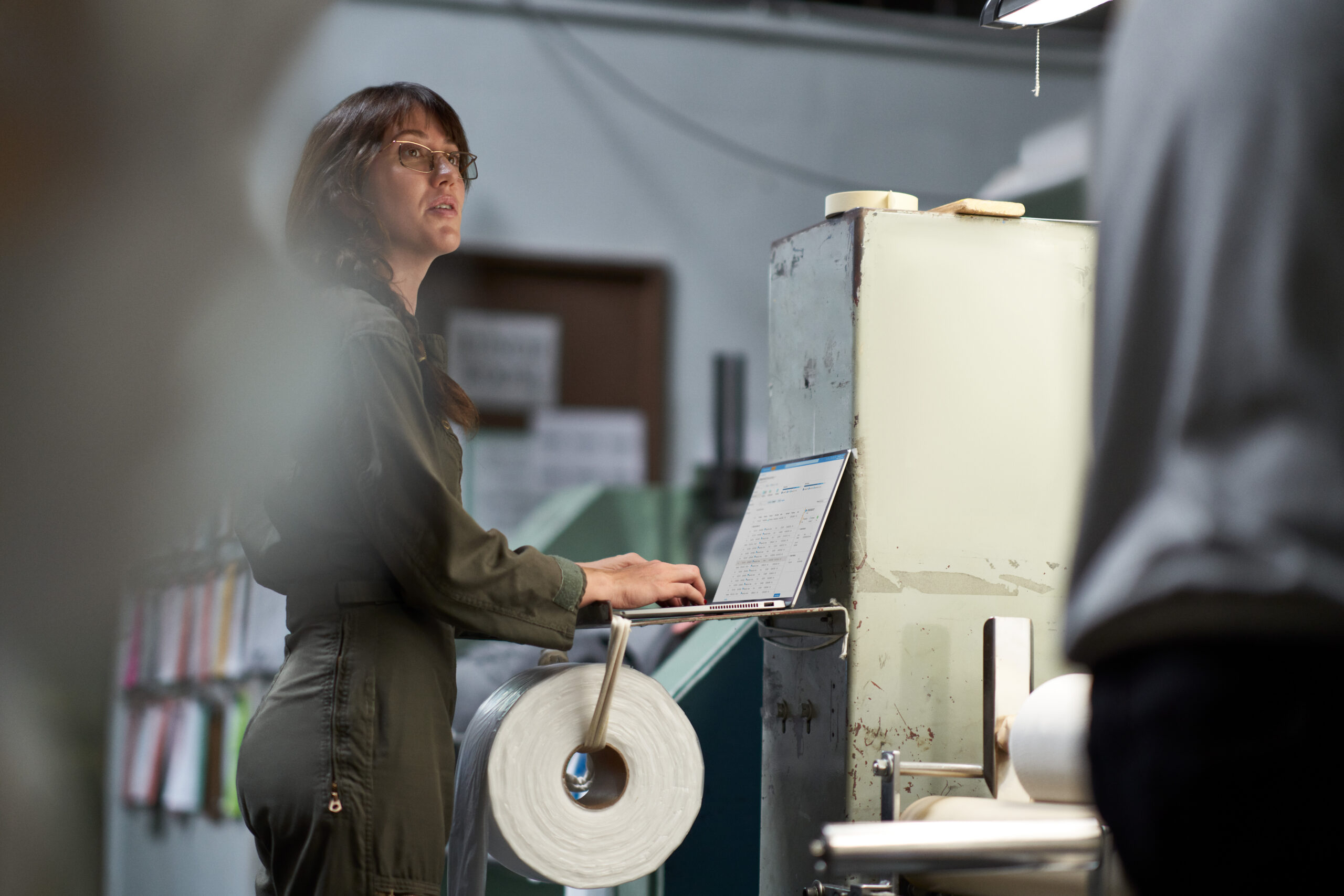 A person standing at an industrial machine, using a laptop while a roll of material is mounted on the equipment in a factory setting.